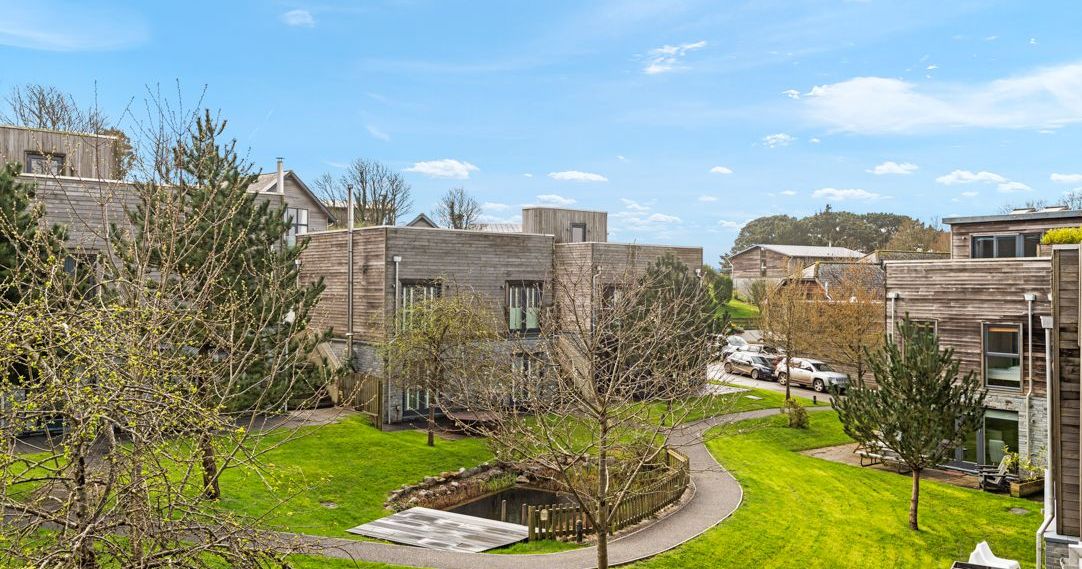 Modern residential buildings with wooden facades, green grass, and a small pond with a pathway under a blue sky.