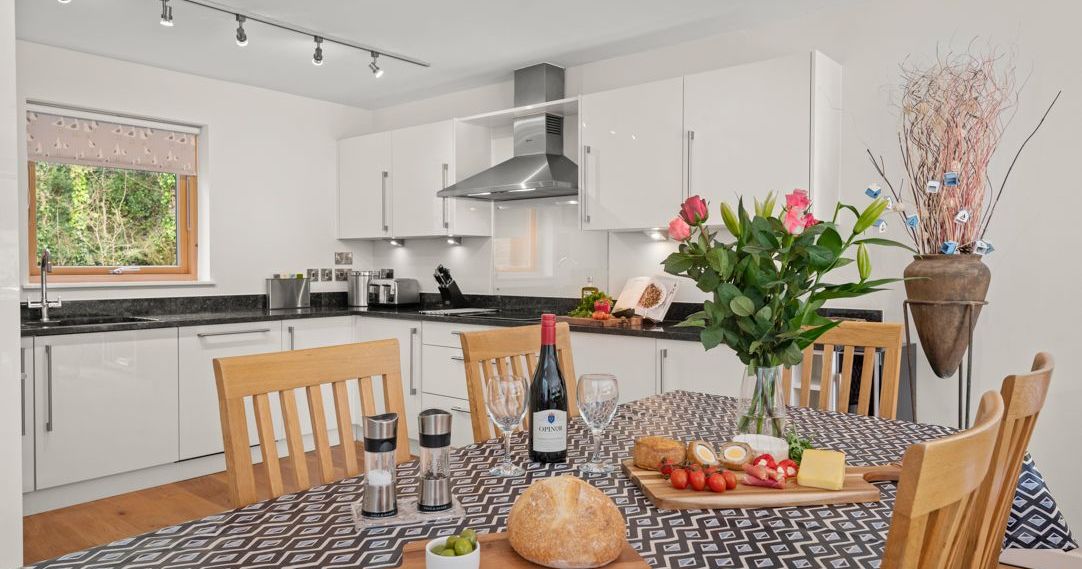 Modern kitchen and dining area with a table set for a meal, including bread, cheese, strawberries, wine, and fresh flowers.