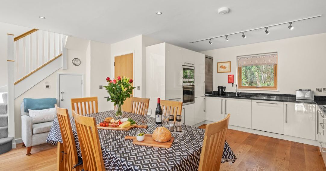 Modern kitchen and dining area with wooden table, six chairs, patterned tablecloth, and food setup