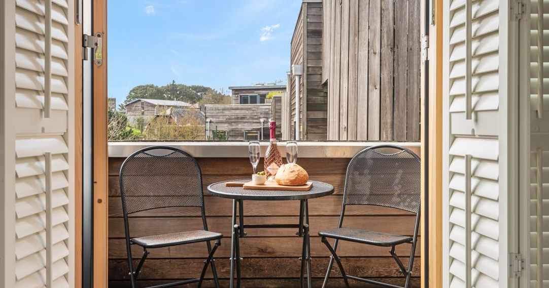 Small balcony with two metal chairs and a round table set with wine, glasses, bread, and cheese.
