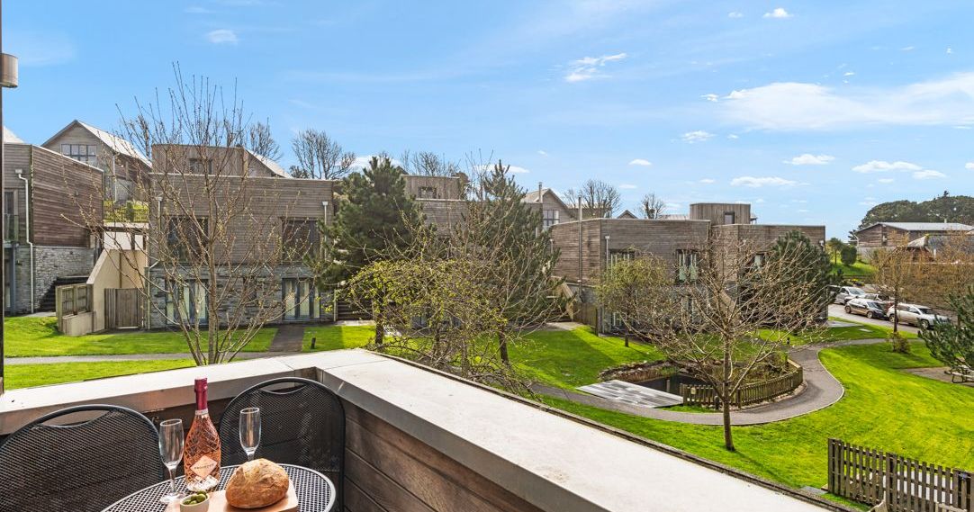 Modern residential complex viewed from a balcony with a small table set for two, featuring bread, olives, a bottle of wine, and glasses.