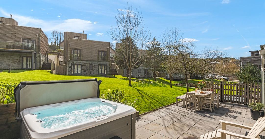 A modern patio with a hot tub, outdoor dining set, and green lawn surrounded by contemporary buildings.