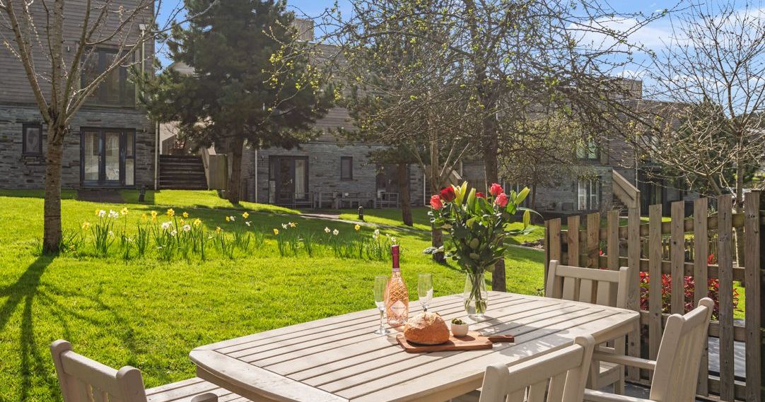 Outdoor wooden table with flowers, bread, and drinks in a garden with modern houses in the background.
