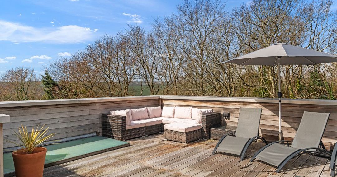 Outdoor wooden deck with patio furniture, sun loungers, umbrella, and scenic tree view