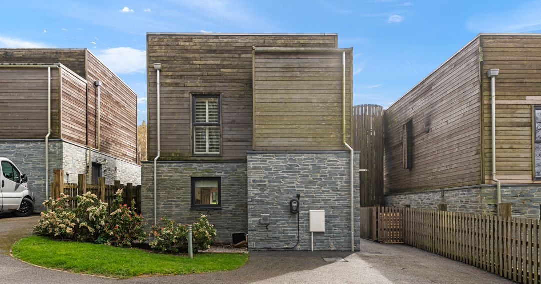 Modern two-story house with wood and stone facade, surrounded by fencing and a small garden.