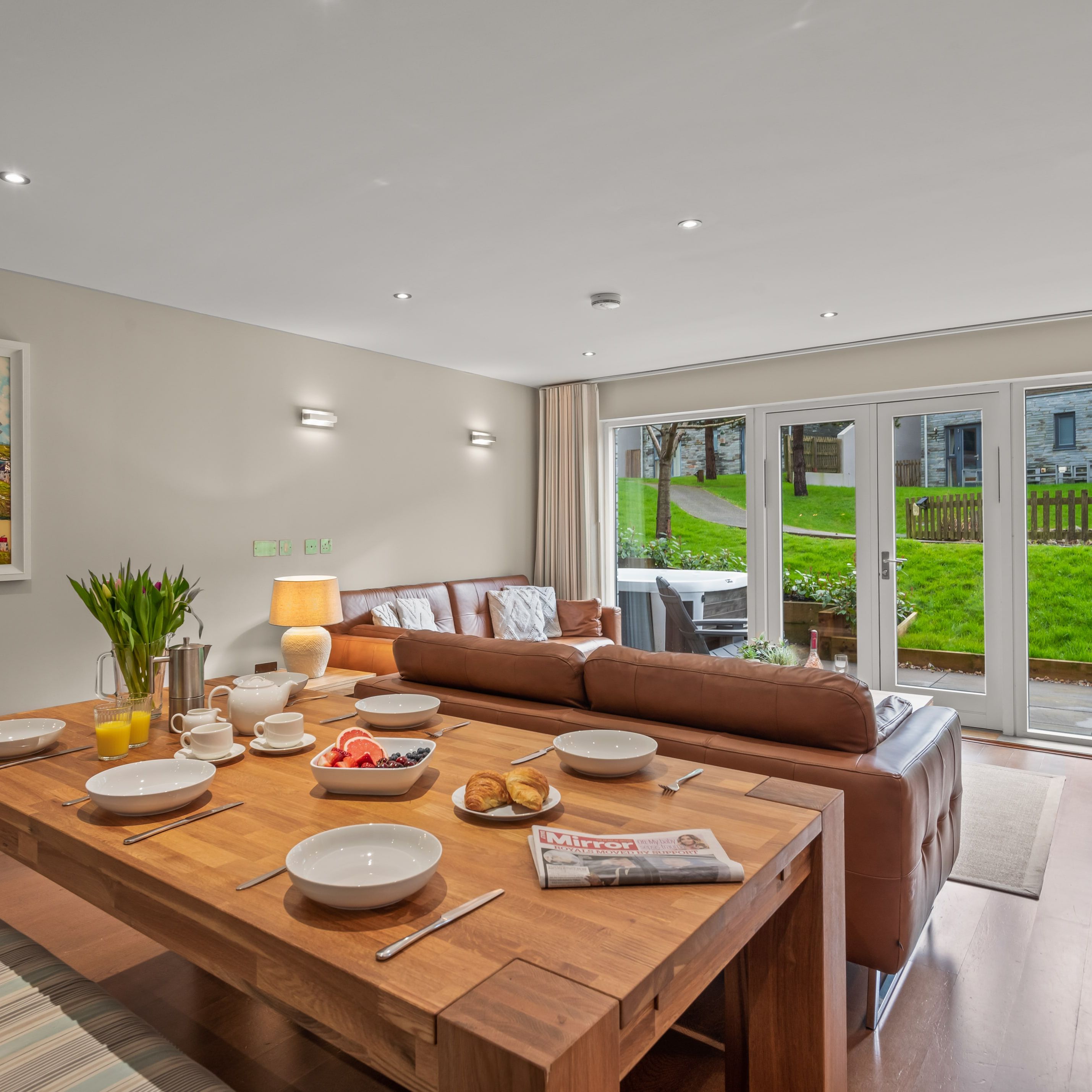 Modern open-plan living and dining area with a wooden table set for breakfast, brown leather sofa, large windows overlooking a green yard, and a colorful beach-themed painting on the wall.