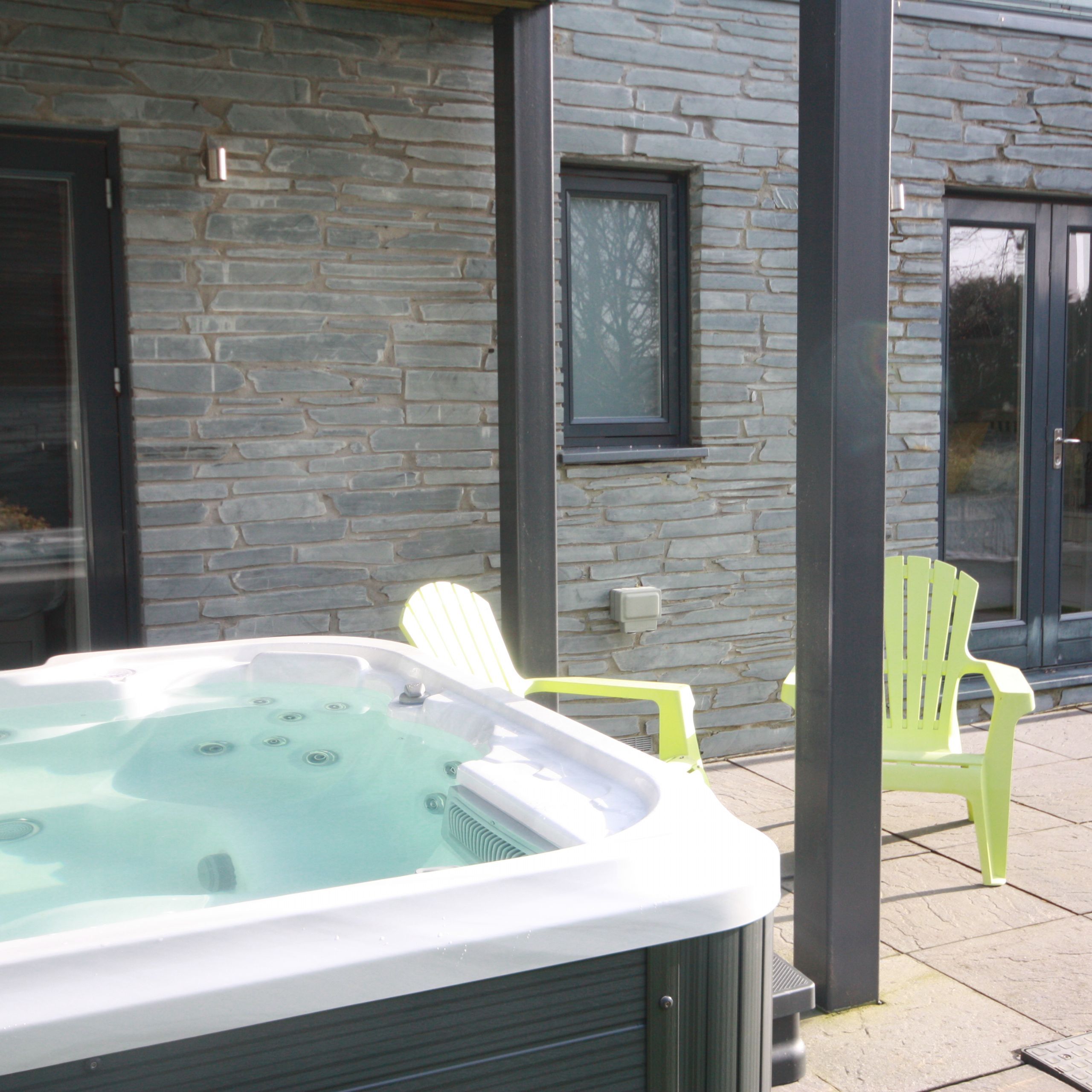 Outdoor hot tub and chairs on a patio next to a modern stone house