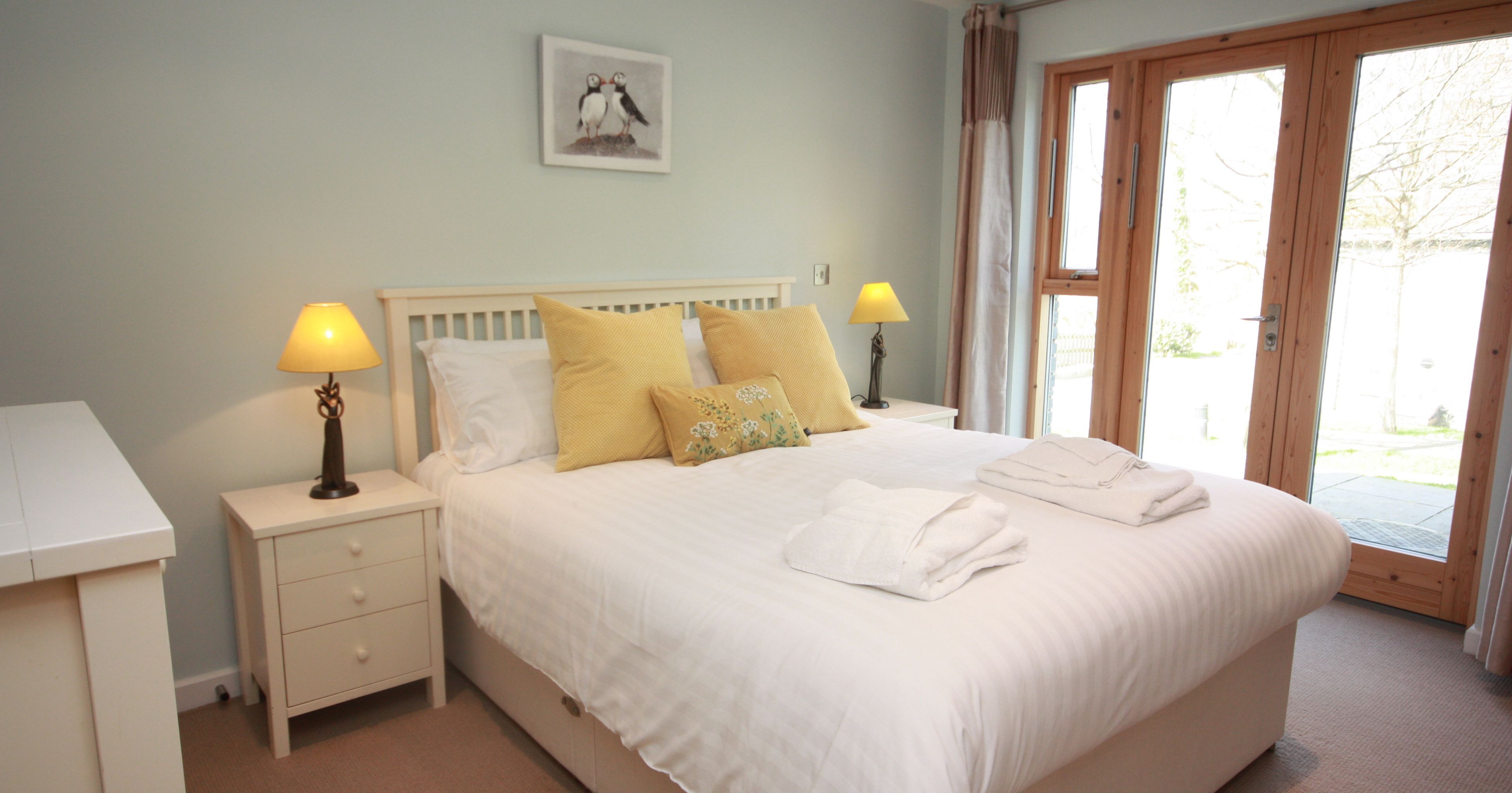 Bright and cozy bedroom with a double bed, white linens, yellow accent pillows, two bedside tables with lamps, and large glass doors letting in natural light.