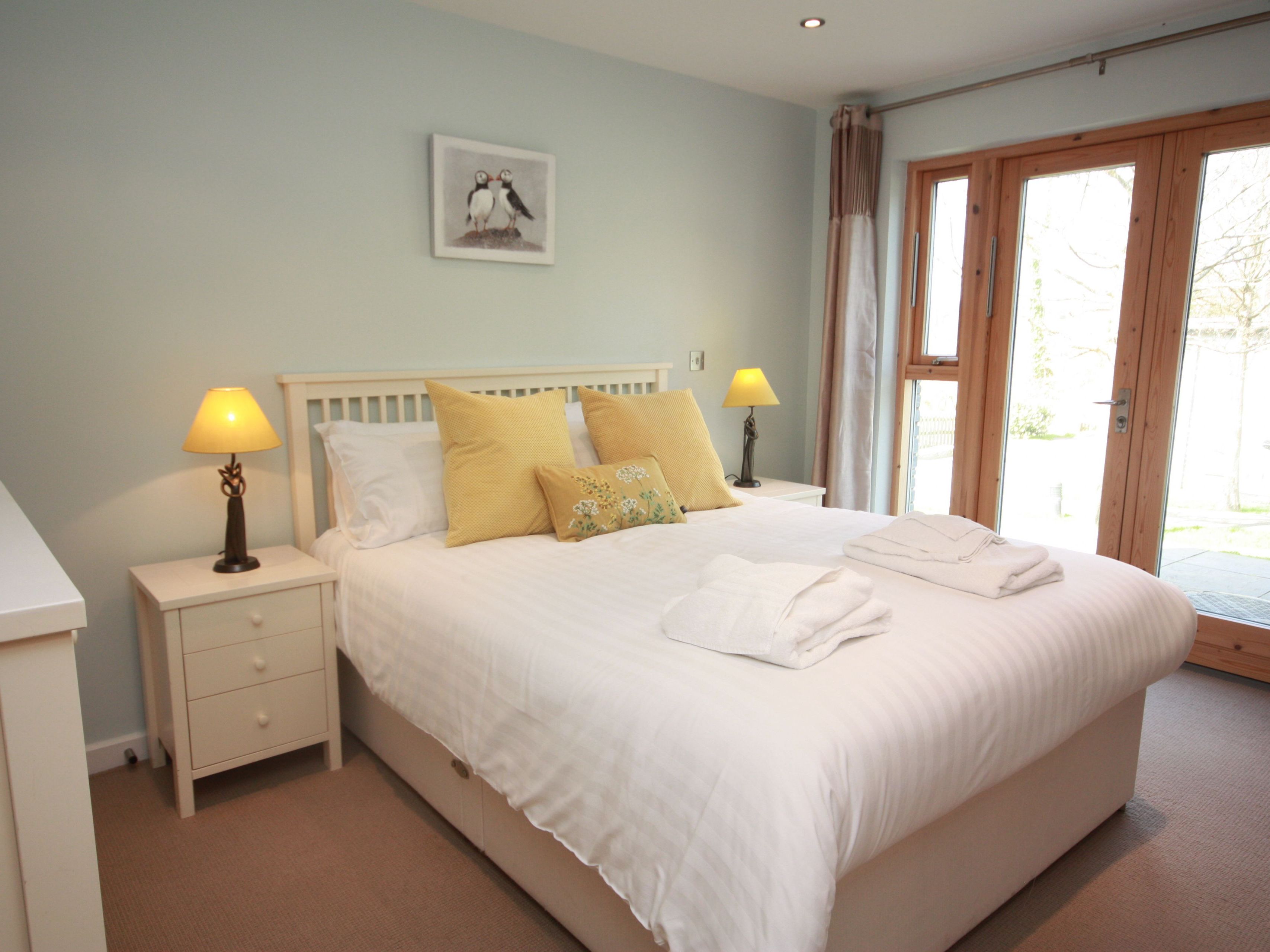 Bright and cozy bedroom with a double bed, white linens, yellow accent pillows, two bedside tables with lamps, and large glass doors letting in natural light.