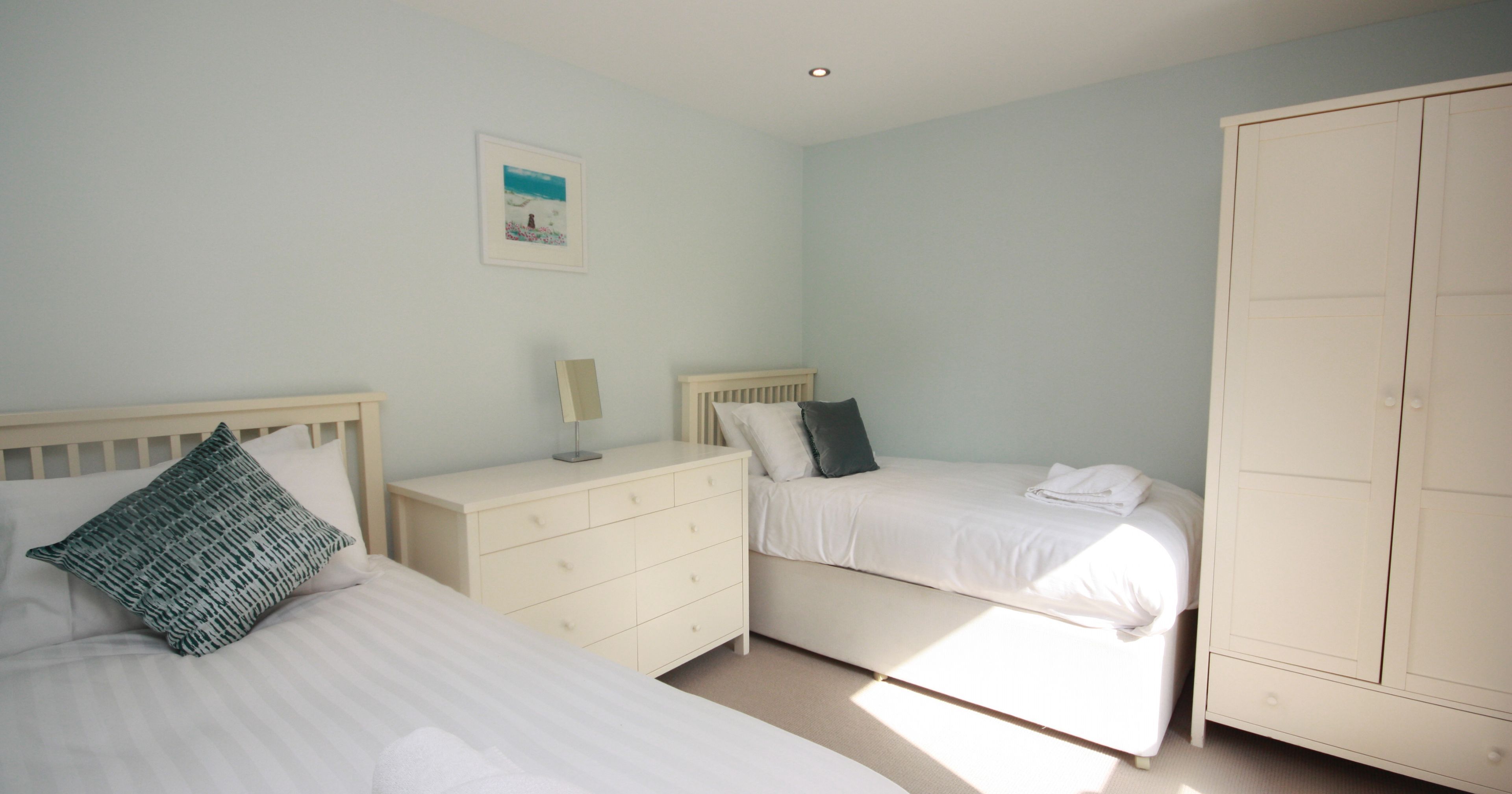 Bright bedroom with two single beds, white furniture, a wardrobe, and a chest of drawers against light blue walls.
