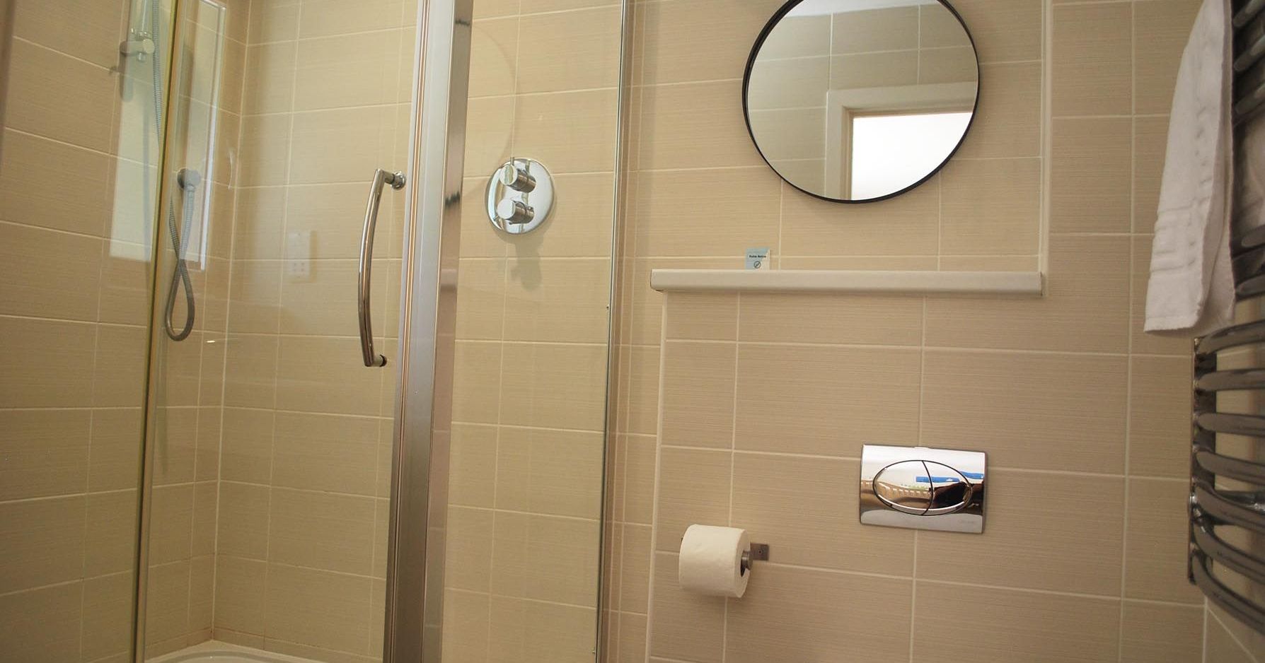 Modern bathroom with glass shower, toilet, round mirror, and beige tiles