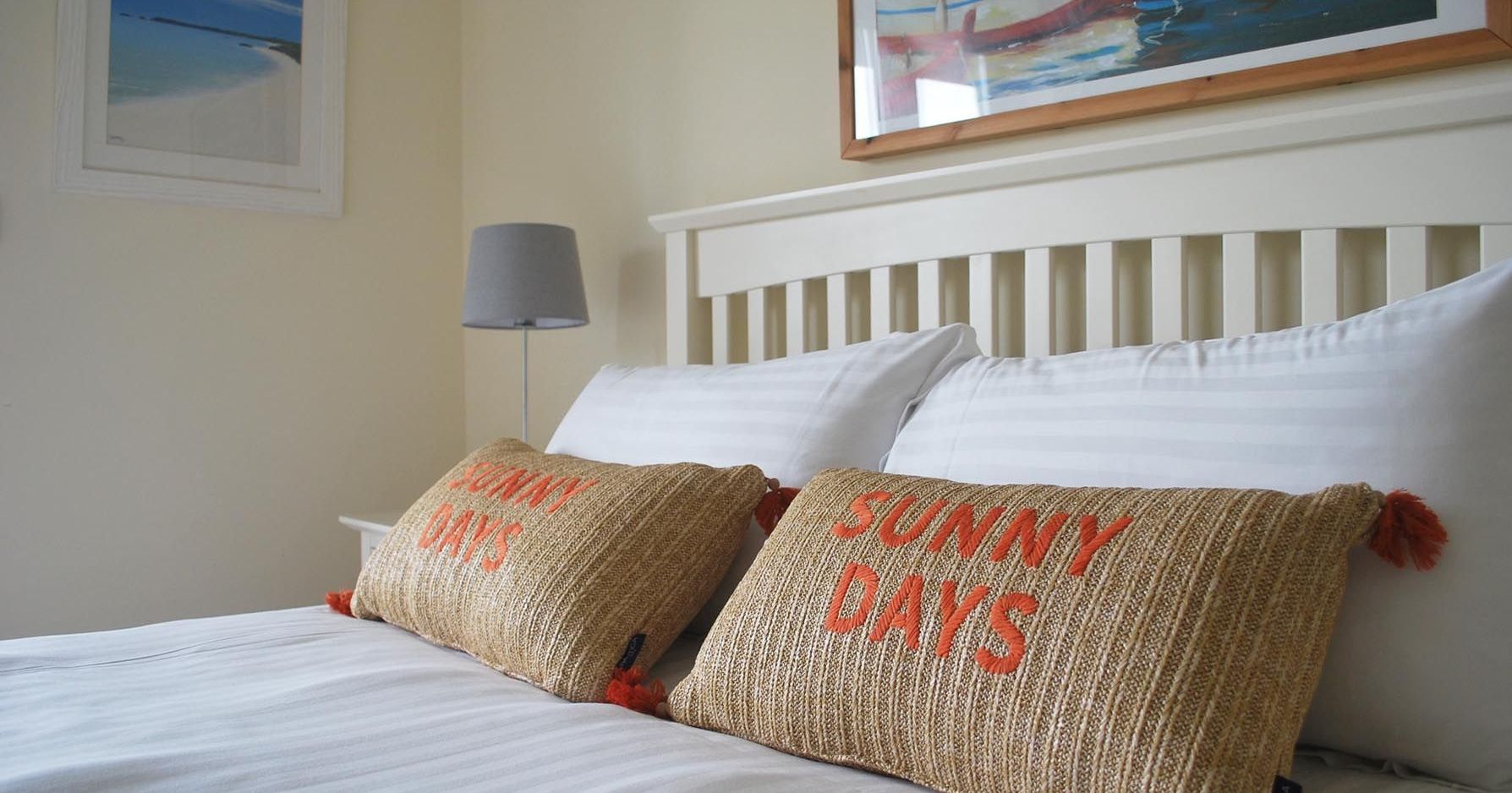 Cozy bed with two decorative pillows labeled 'Sunny Days', white bedding, a lamp, and framed beach and boat pictures on the wall.