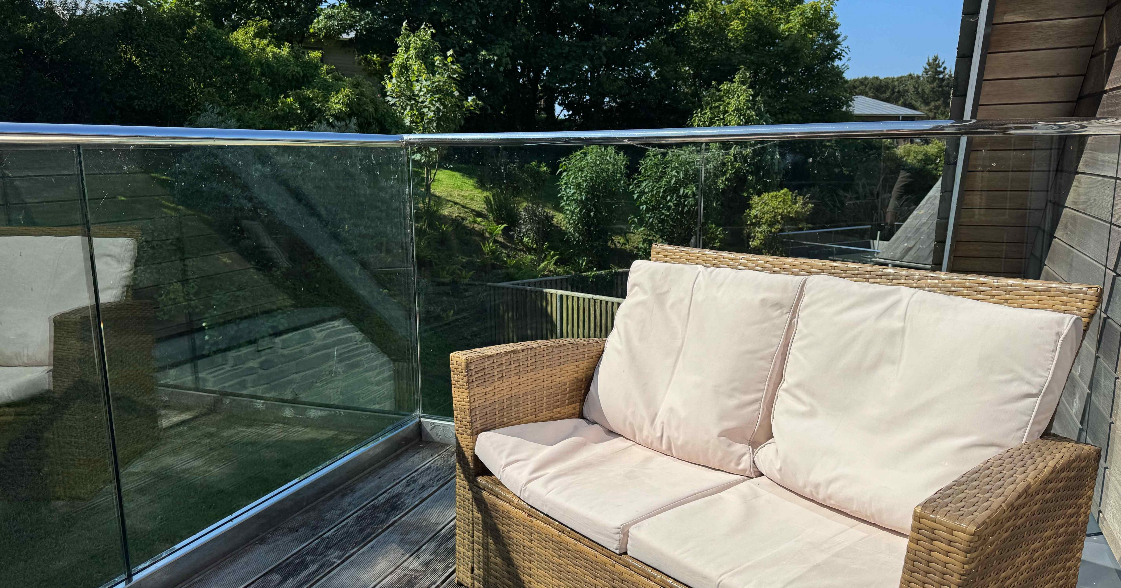 Wicker outdoor loveseat with light-colored cushions on a balcony with glass railing, surrounded by greenery.