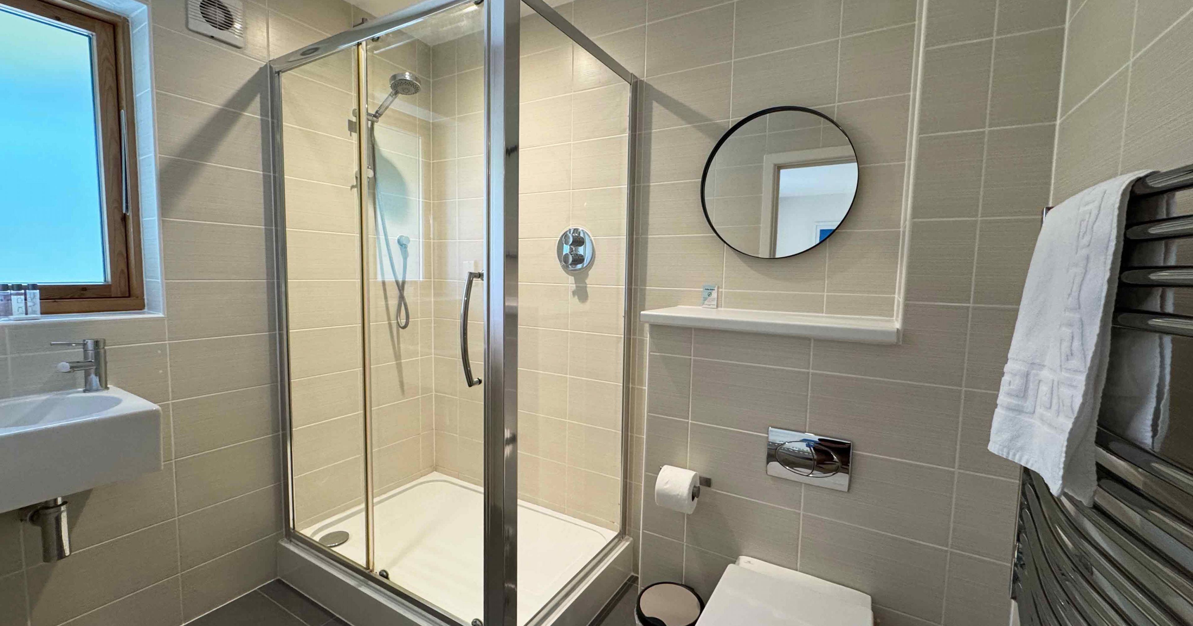 Modern bathroom with glass shower enclosure, toilet, round mirror, and towel rail.