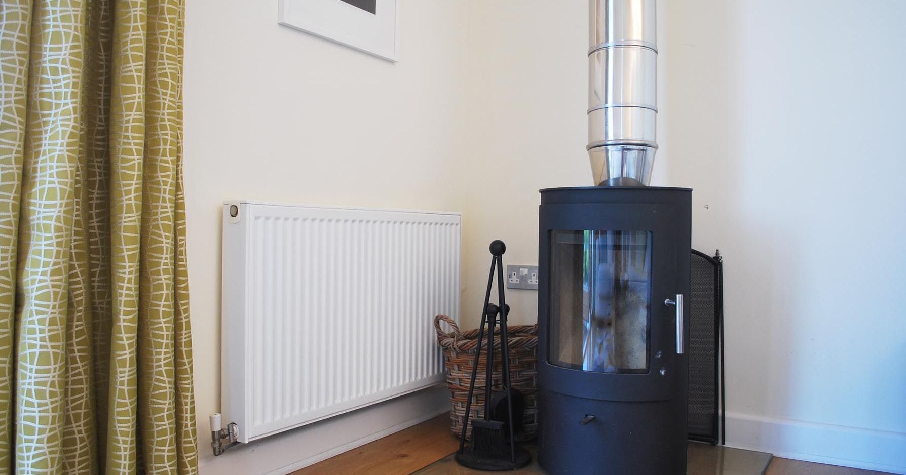Modern wood burning stove with metal chimney next to a radiator and a basket in a corner of a room