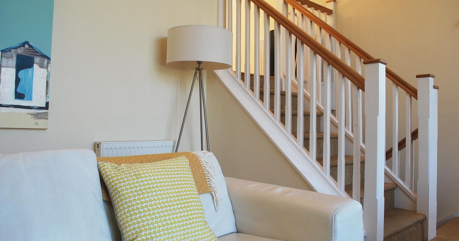 Cozy living room with a beige sofa, colorful cushions, a floor lamp, and a staircase with white railings and a wooden handrail.