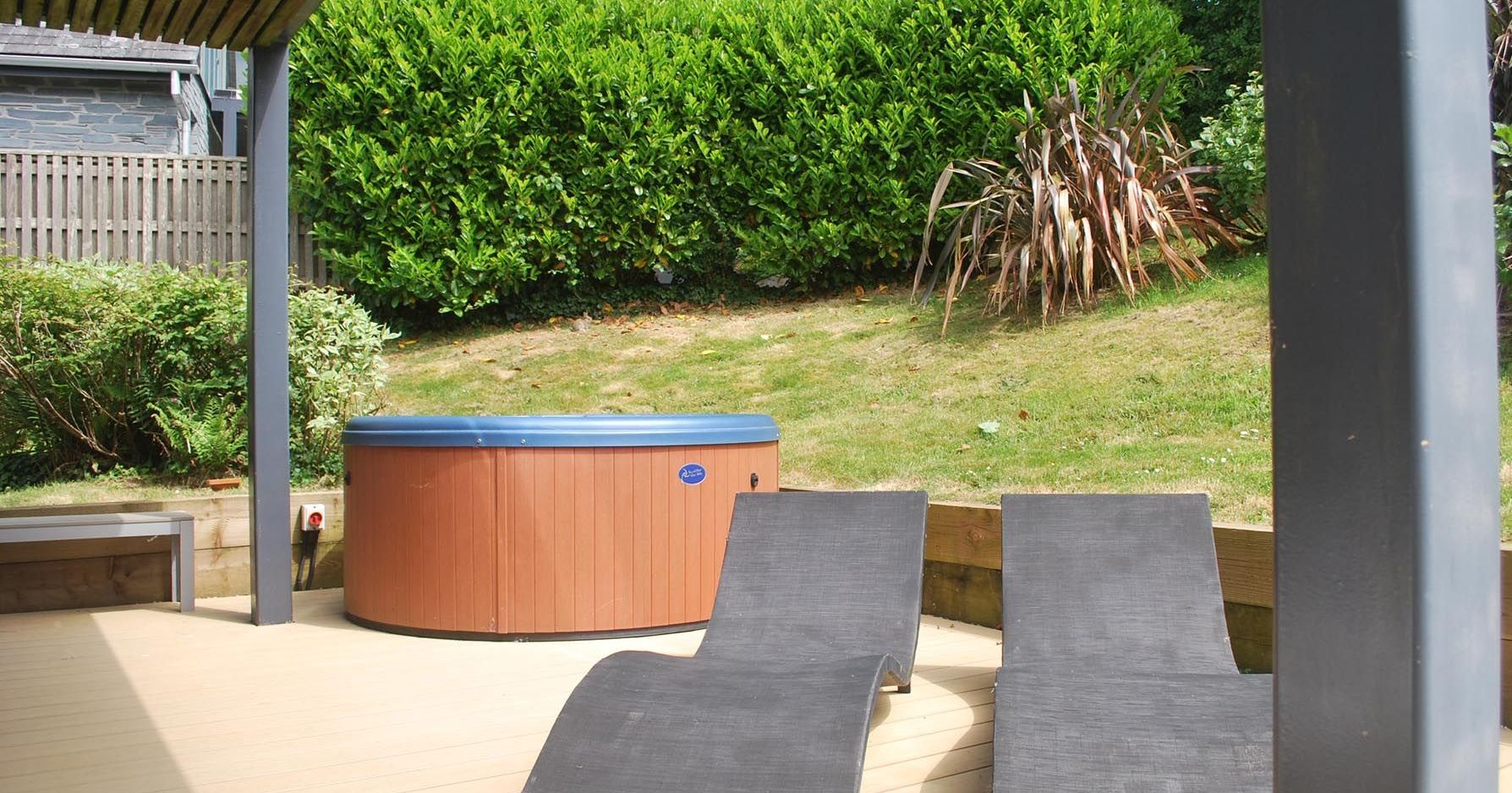 Outdoor patio area with two black lounge chairs and a hot tub against a backdrop of green bushes.