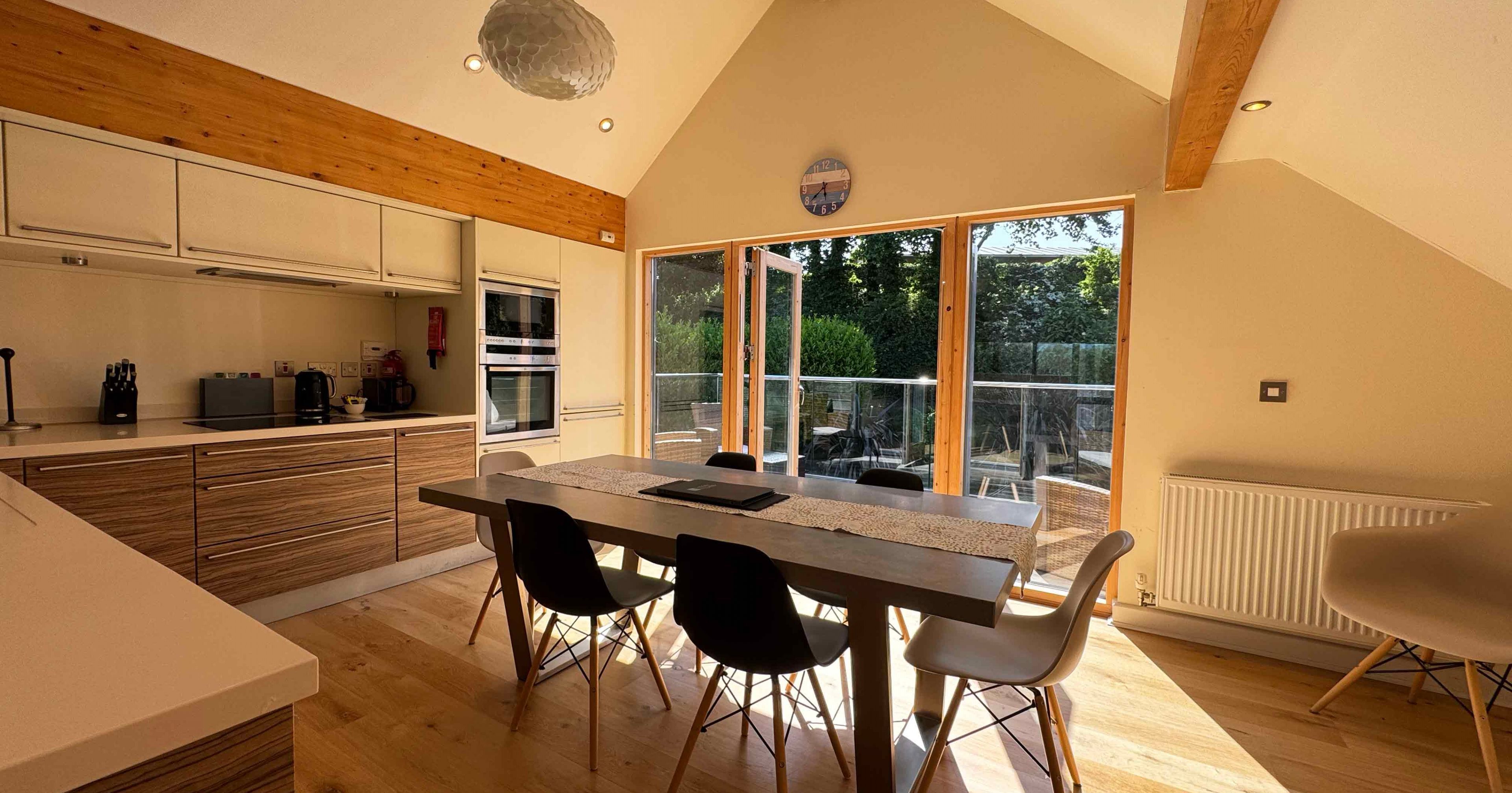 Modern kitchen and dining area with wooden beams, large table and chairs, and wide glass doors opening to a patio.
