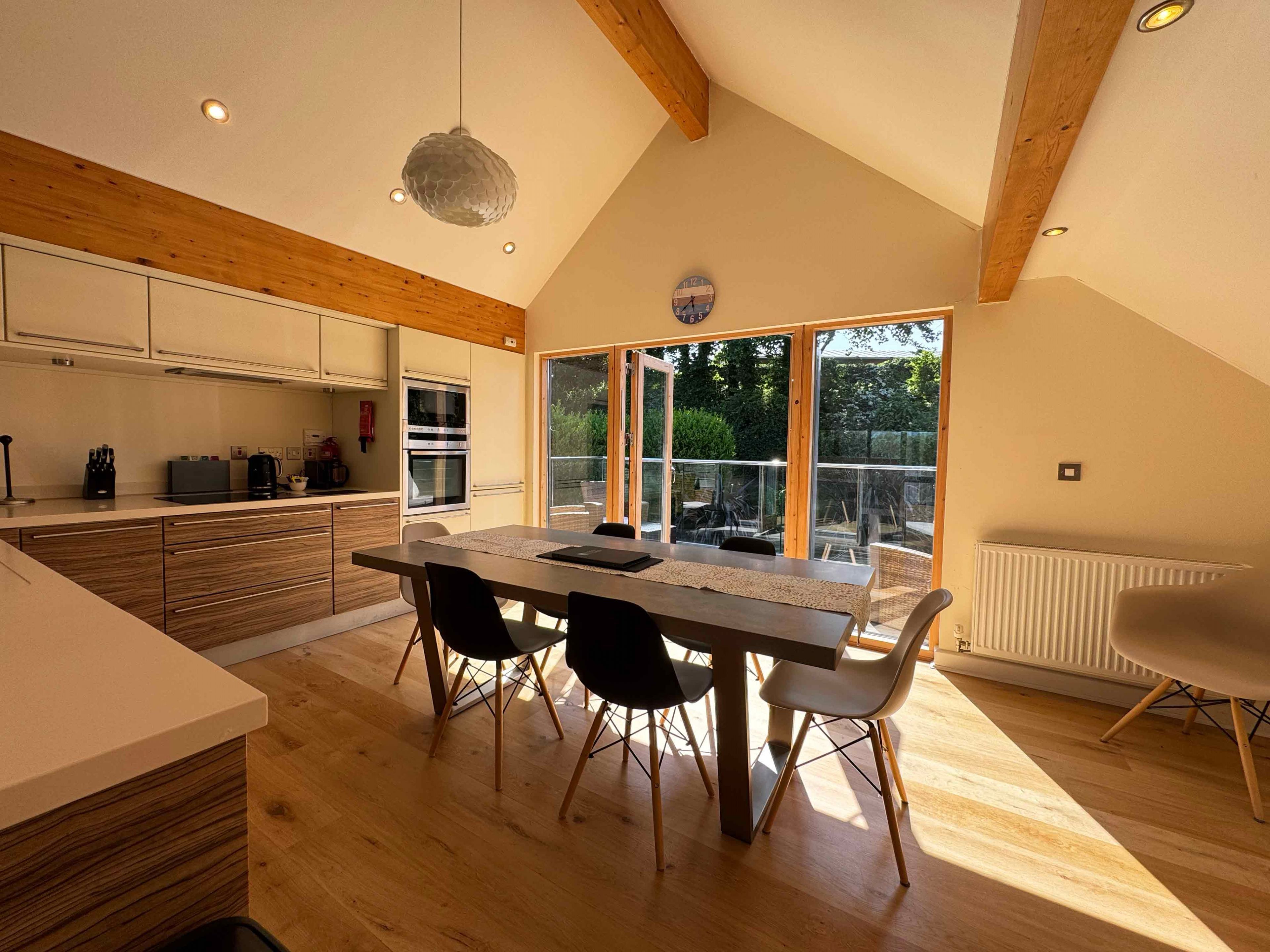 Modern kitchen and dining area with wooden beams, large table and chairs, and wide glass doors opening to a patio.