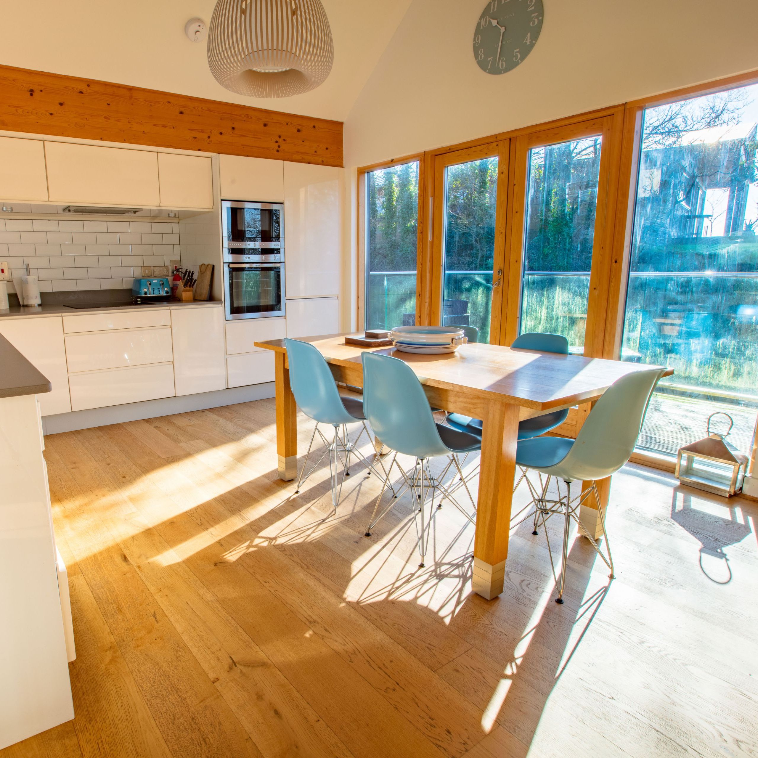 Modern kitchen and dining area with wooden floor, white cabinets, blue chairs, and large windows letting in sunlight.