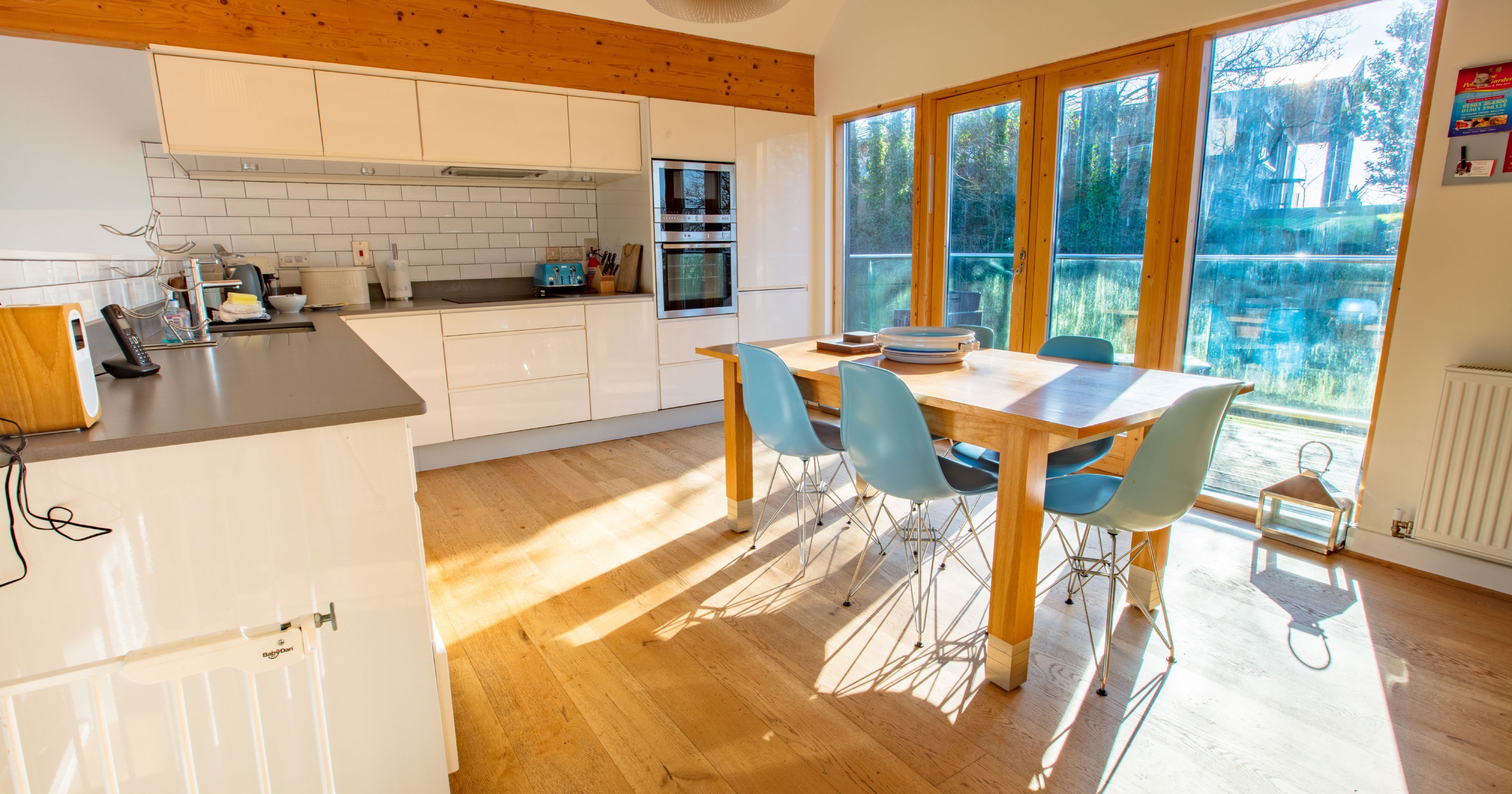 Modern kitchen and dining area with wooden floor, white cabinets, blue chairs, and large windows letting in sunlight.