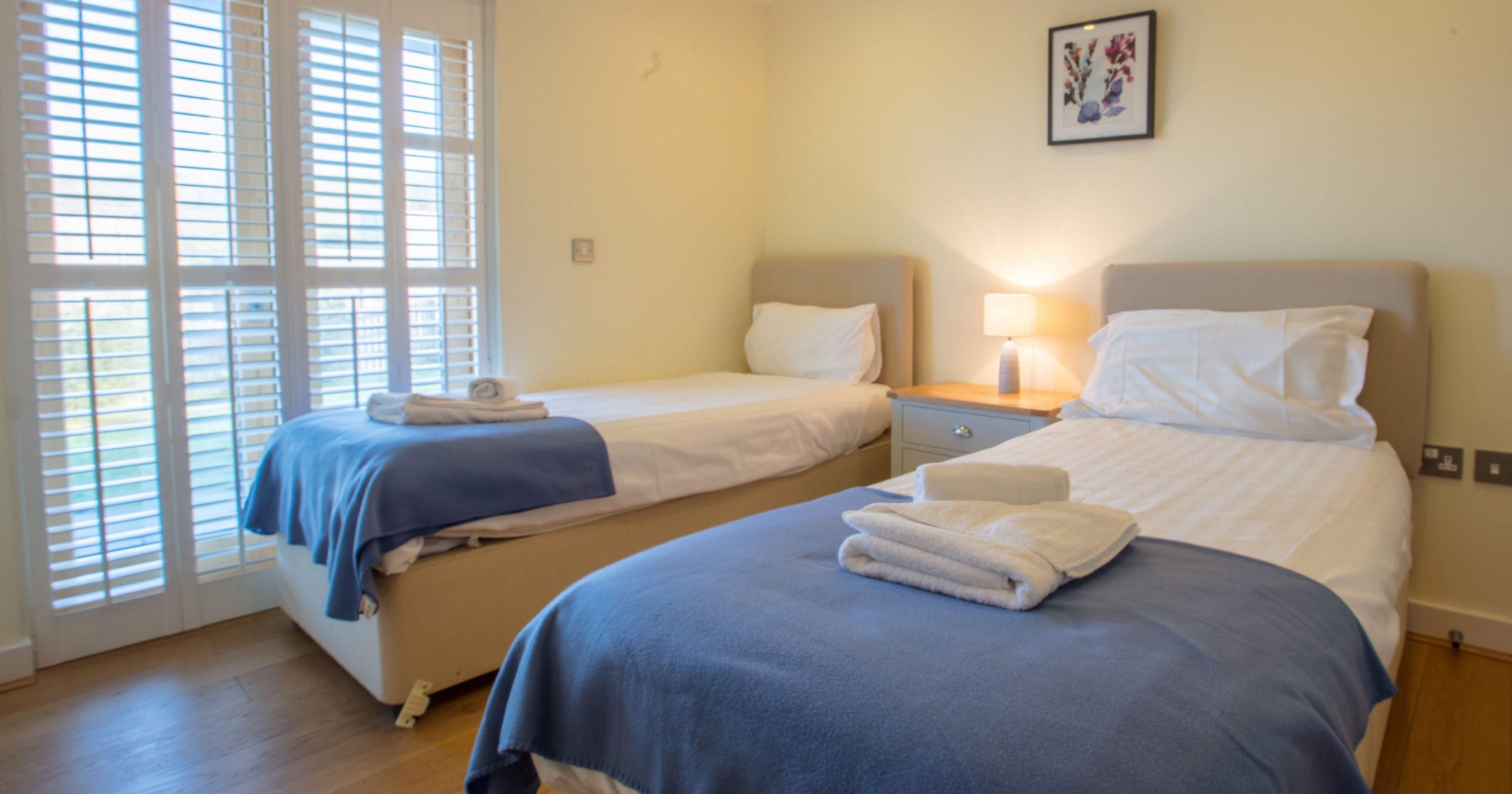 Two single beds with blue blankets in a bright bedroom with wooden floor and white shutters.