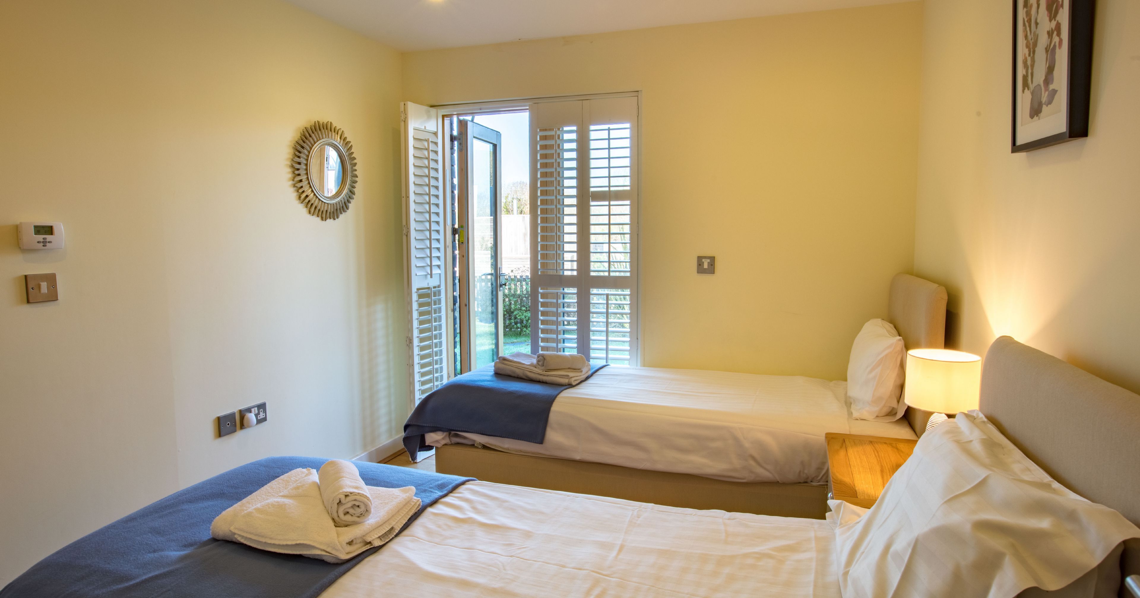 Bright bedroom with two single beds, white linens, a nightstand with a lamp, an open door with shutters, and wall art.