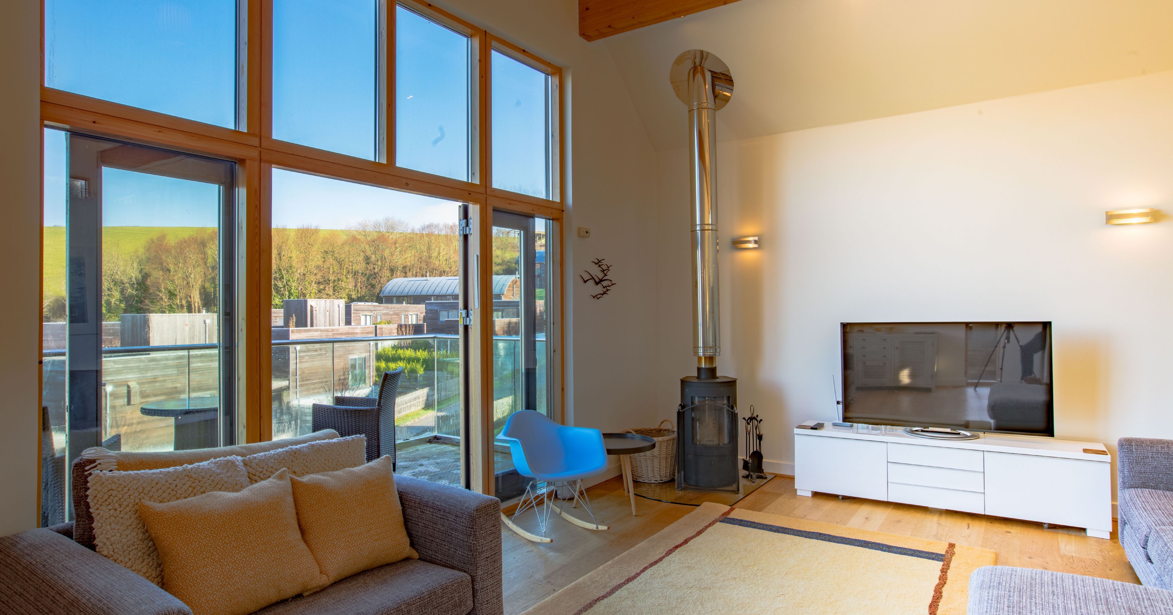 Modern living room with large glass windows, a gray sofa, TV, wood stove, and access to a balcony