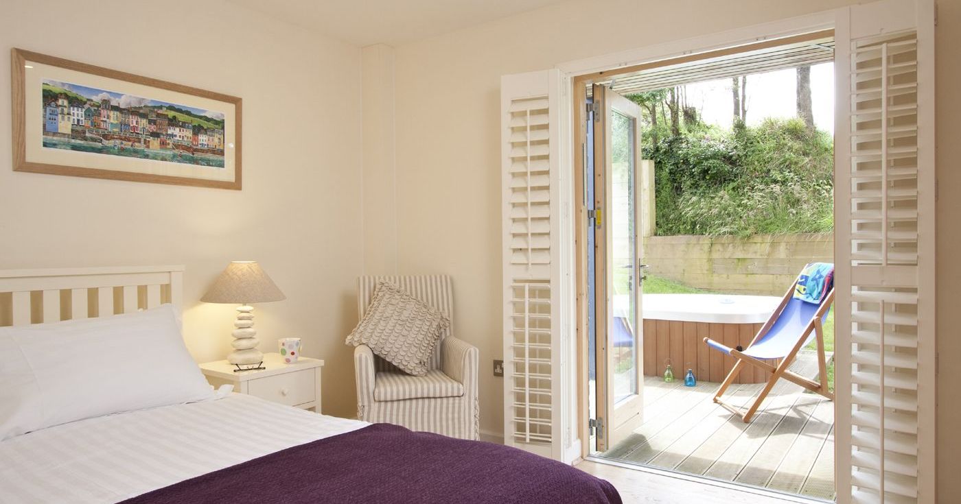 Bright bedroom with a double bed, purple blanket, and open French doors leading to a patio with a deck chair and hot tub.