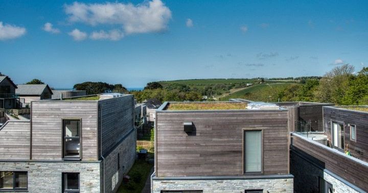 Modern wooden houses with flat roofs against a backdrop of green hills and a blue sky with clouds.