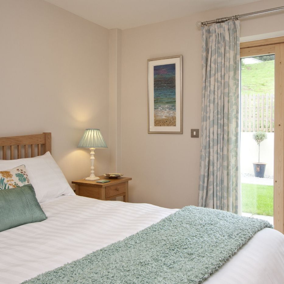 Cozy bedroom with wooden bed, white linens, green accents, nightstand with lamp, and large glass doors to a garden.