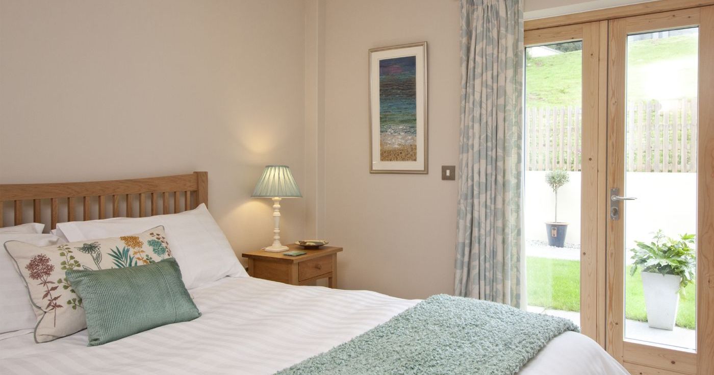 Cozy bedroom with wooden bed, white linens, green accents, nightstand with lamp, and large glass doors to a garden.