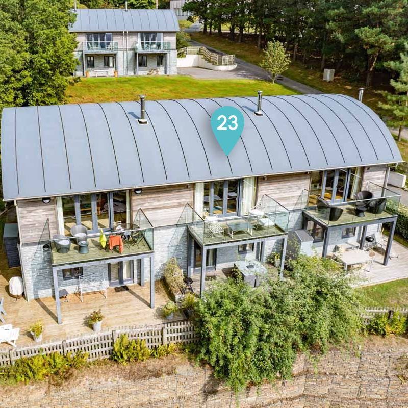 Modern multi-storey holiday lodge with balconies, outdoor seating, and surrounded by trees, marked with number 23.