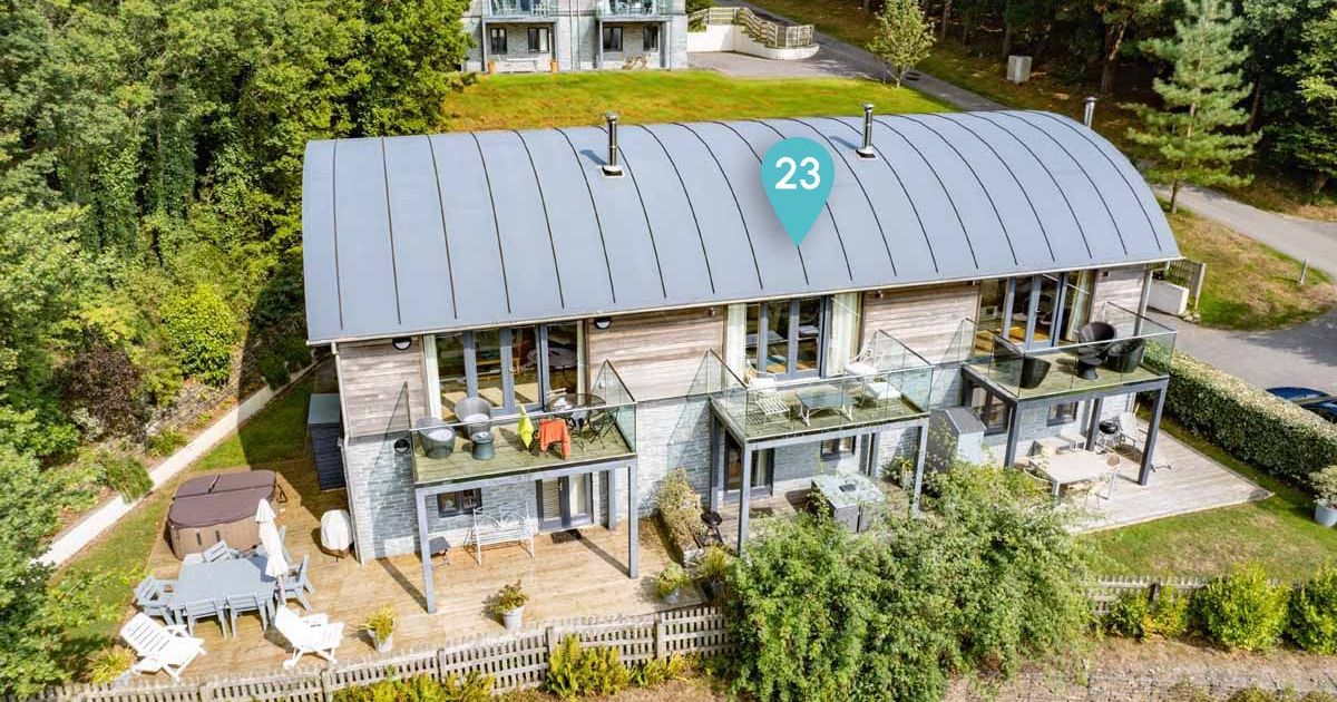 Modern multi-storey holiday lodge with balconies, outdoor seating, and surrounded by trees, marked with number 23.
