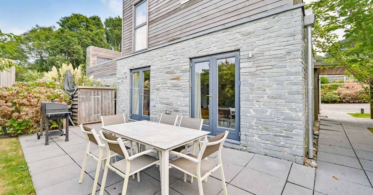 Modern outdoor patio with dining table, chairs, and a barbecue grill next to a stone and wood house.