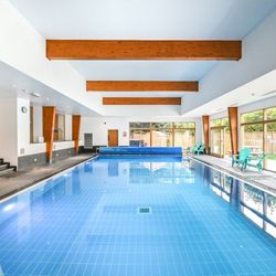 Indoor swimming pool with blue tiled floor, wooden ceiling beams, large windows, and poolside seating.