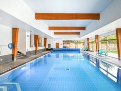 Indoor swimming pool with blue tiled floor, wooden ceiling beams, large windows, and poolside seating.