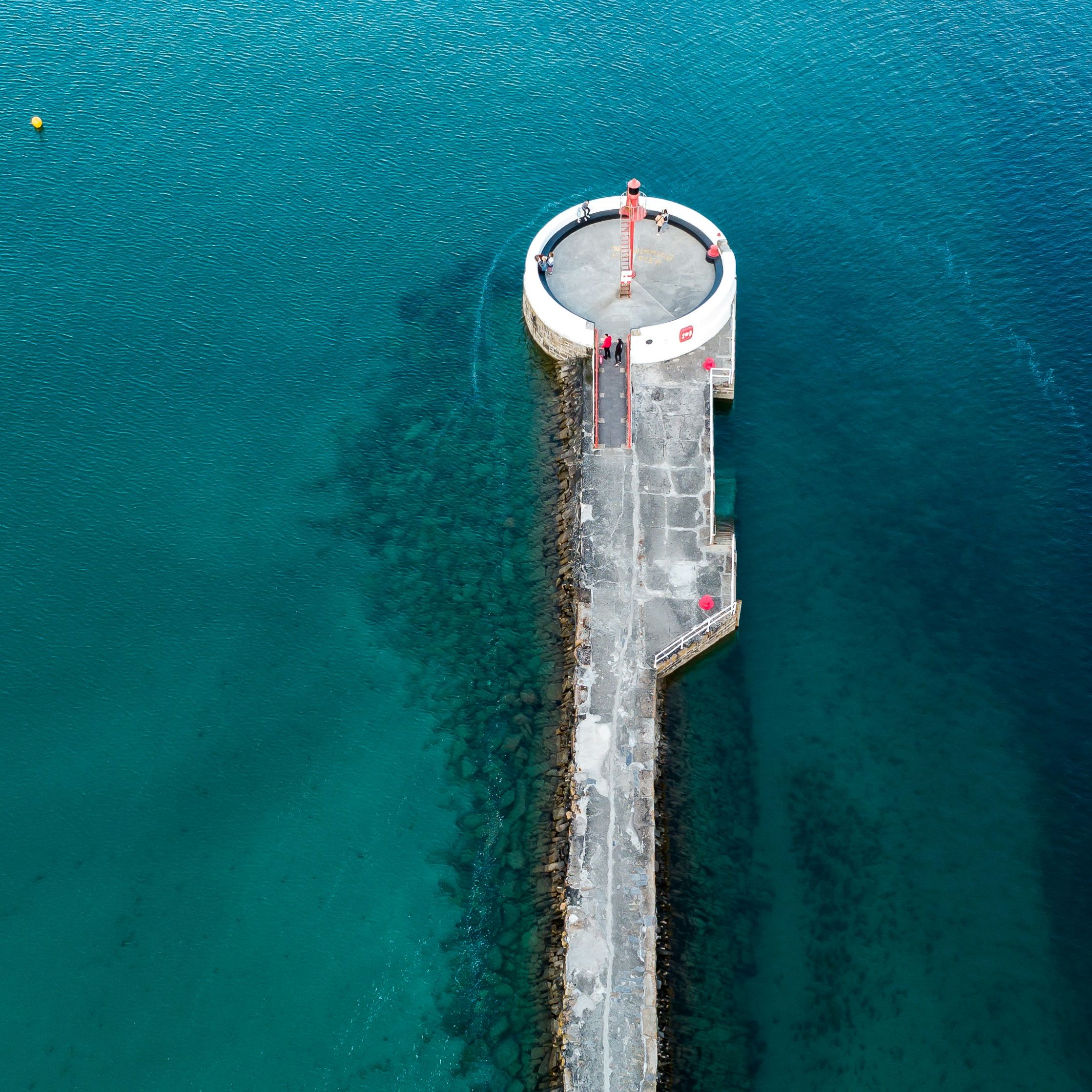 Aerial view of a circular lighthouse at the end of a long pier extending into clear blue water