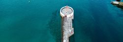 Aerial view of a circular lighthouse at the end of a long pier extending into clear blue water