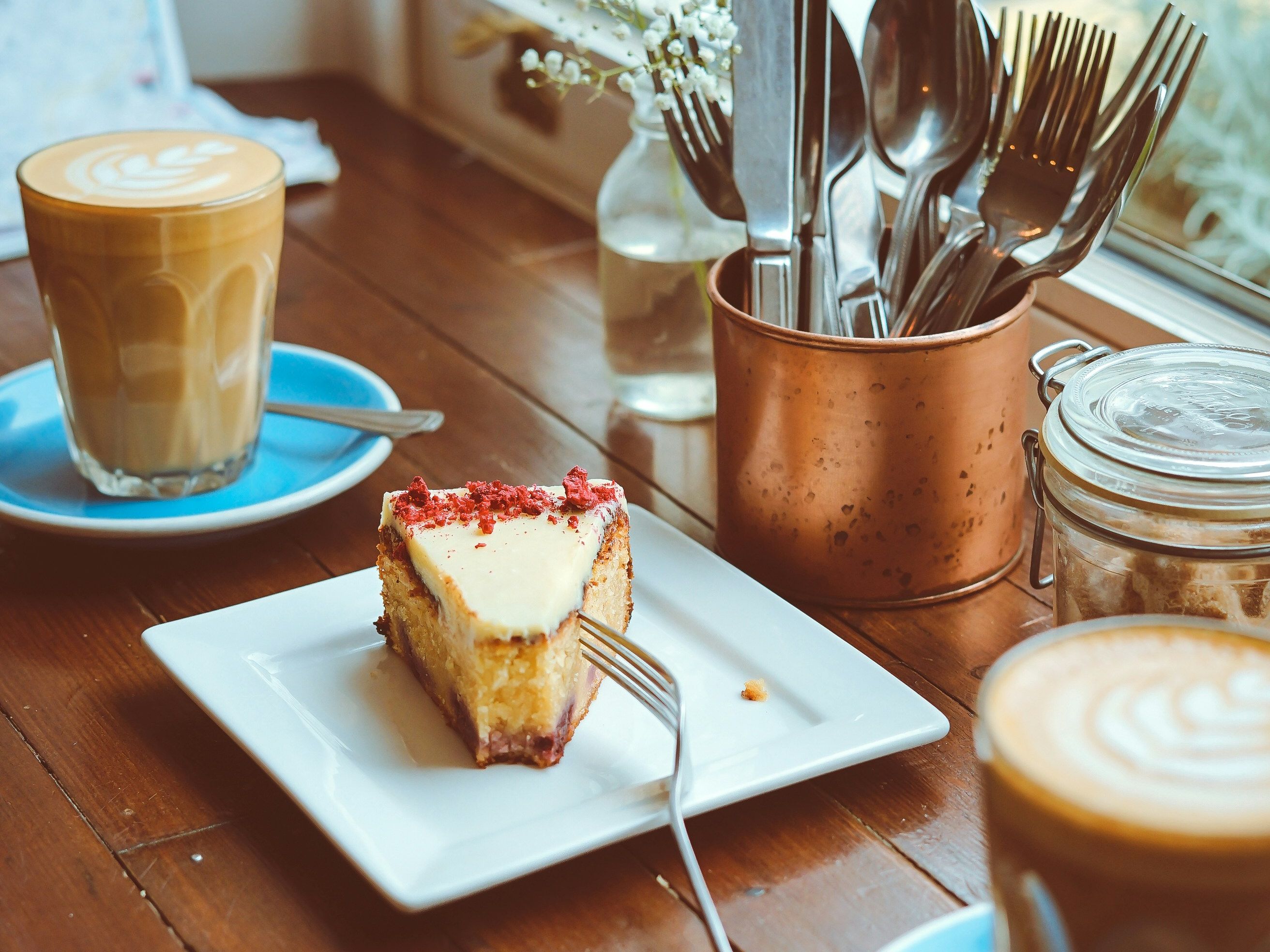 A slice of cake on a plate with two cups of latte art coffee on a wooden table by a window.