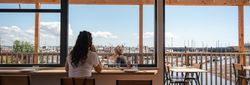 Person sitting at a cafe window with a view of a marina and boats outside.