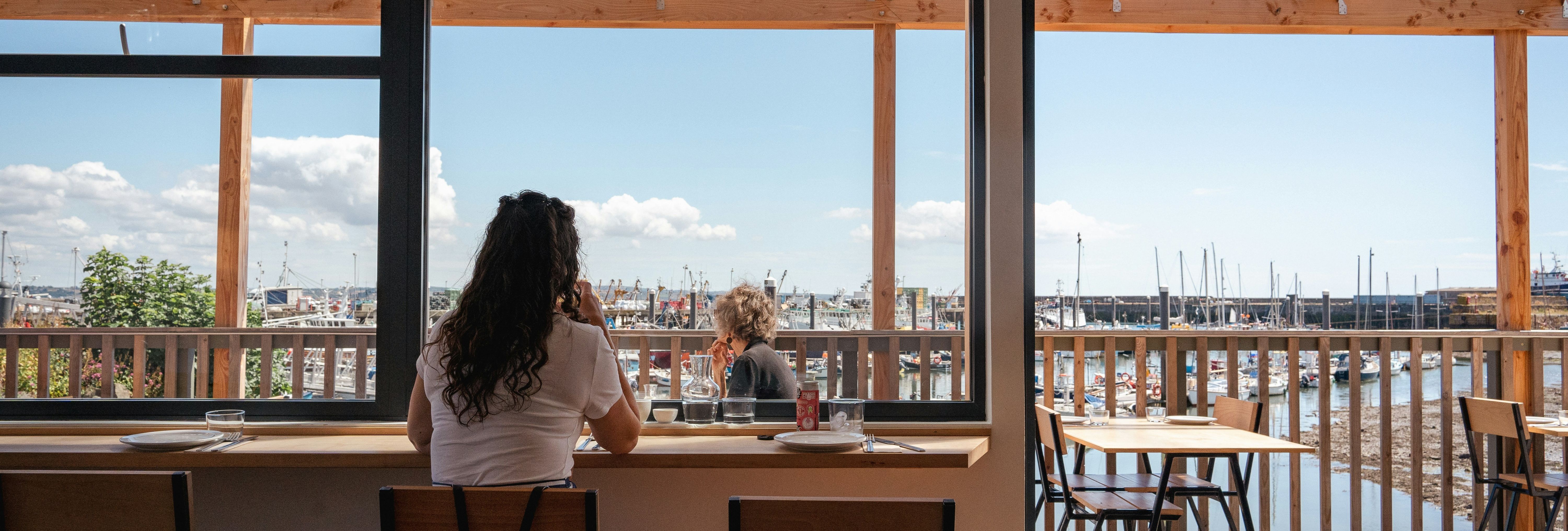 Person sitting at a cafe window with a view of a marina and boats outside.