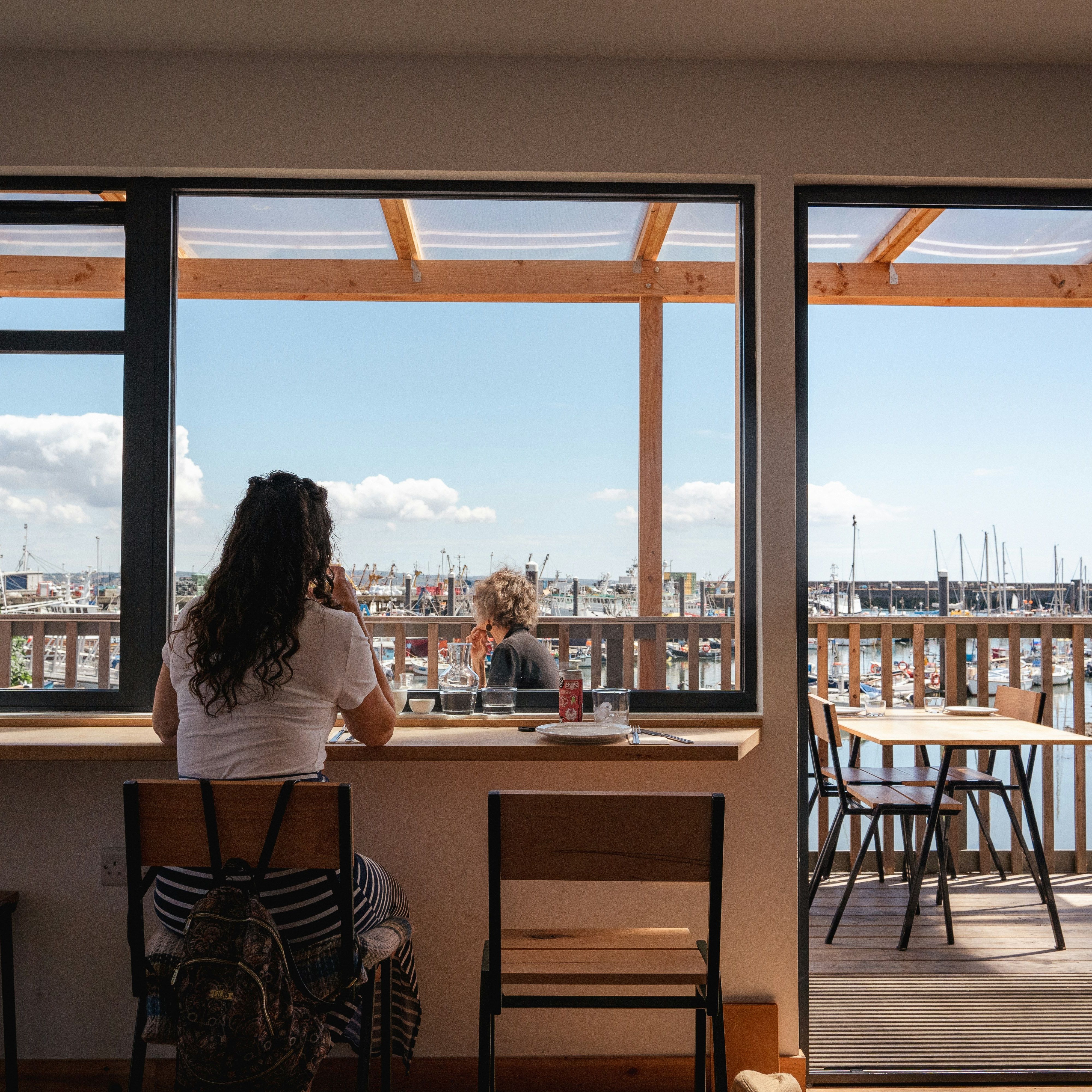 Person sitting at a cafe window with a view of a marina and boats outside.