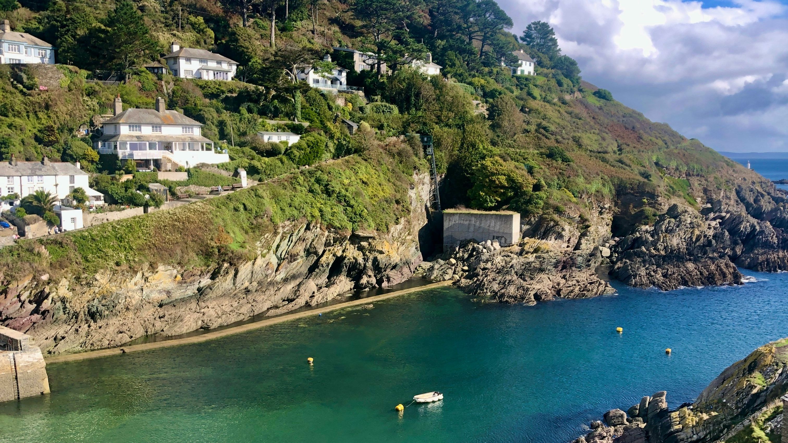 Scenic view of houses on a lush green hillside overlooking a calm turquoise bay with rocky cliffs and clear skies.
