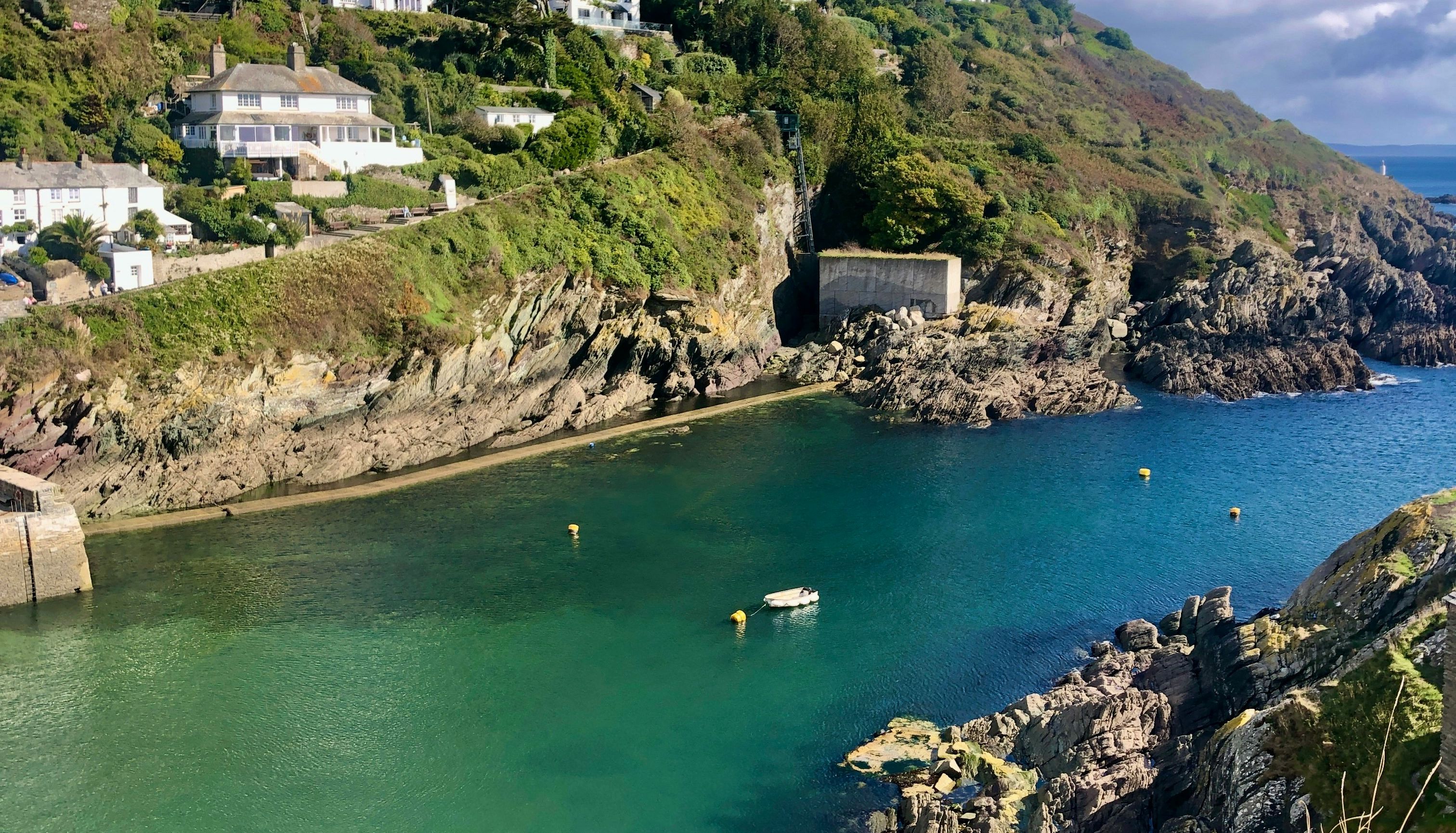 Scenic view of houses on a lush green hillside overlooking a calm turquoise bay with rocky cliffs and clear skies.