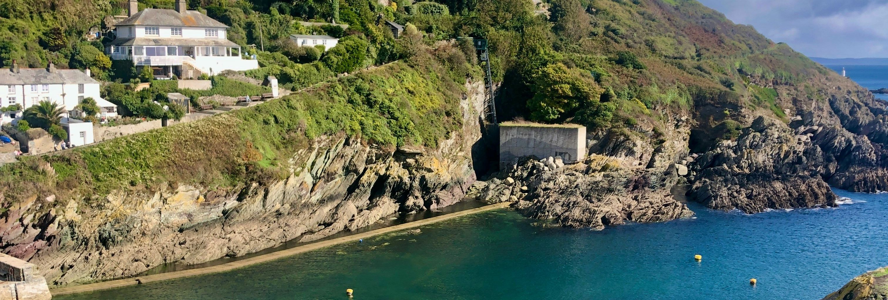 Scenic view of houses on a lush green hillside overlooking a calm turquoise bay with rocky cliffs and clear skies.