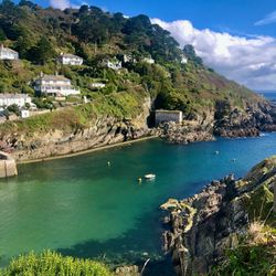 Scenic view of houses on a lush green hillside overlooking a calm turquoise bay with rocky cliffs and clear skies.
