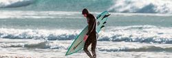 Surfer in a wetsuit carrying a surfboard on a beach with waves in the background
