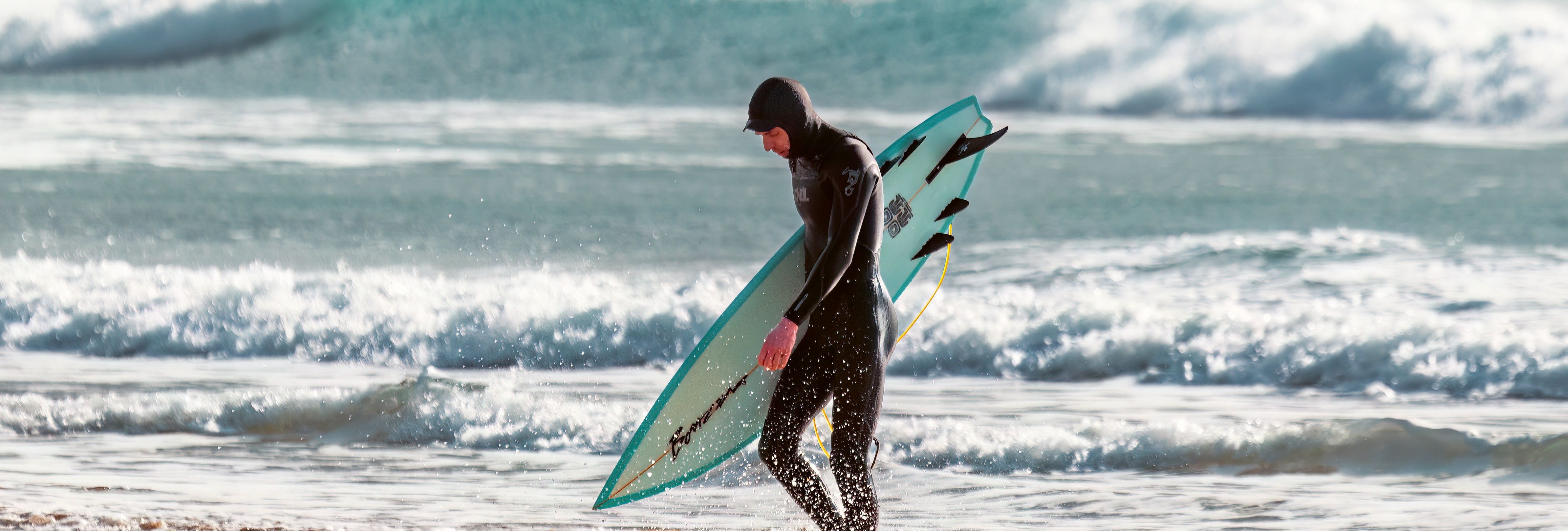 Surfer in a wetsuit carrying a surfboard on a beach with waves in the background