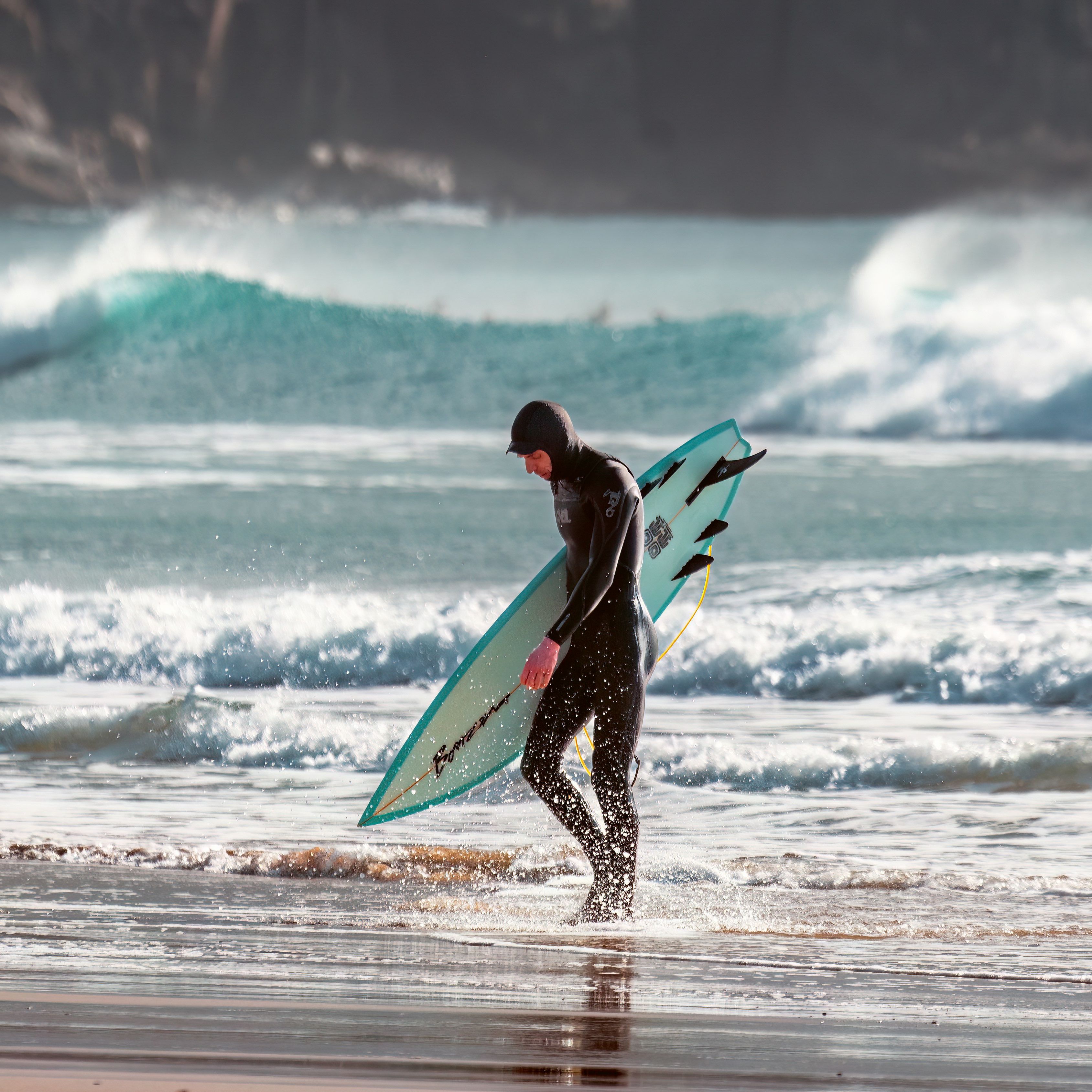 Surfer in a wetsuit carrying a surfboard on a beach with waves in the background